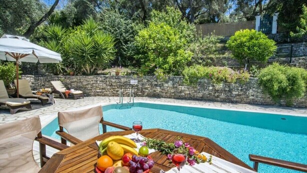 Un área junto a la piscina con una mesa dispuesta con frutas y una copa de vino tinto, rodeada de sillones de descanso y un muro de piedra.