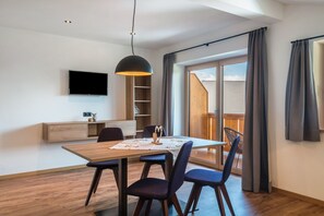Ein moderner Essbereich mit einem Holztisch, dunklen Stühlen, einer Pendelleuchte, einem Flachbildfernseher und einer Glastür zum Balkon.