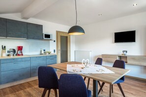 Uma cozinha moderna com armários cinza, uma mesa de jantar de madeira com cadeiras azuis, uma luminária pendente e uma televisão de tela plana fixada na parede.
