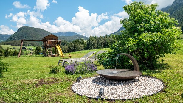 Una cabaña de madera, un parque infantil con columpios, una fuente de piedra circular y un paisaje verde exuberante con montañas al fondo.