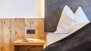 Una mesita de noche de madera con un libro, un enchufe y una cama con almohadas.