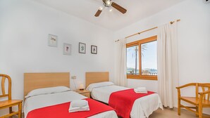 A hotel room with two beds, a window with a view, a ceiling fan, and a wooden chair. - 'Pine Walk' Apartment with Sea Views, Wi-Fi and Air Conditioning (Pollensa)
