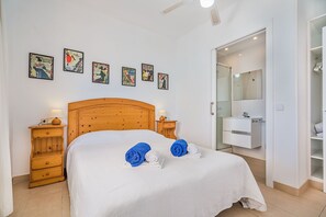 Un dormitorio con cabecera de madera, cama con ropa blanca, dos mesitas de noche con lámparas, un armario y un baño con espejo y lavamanos.