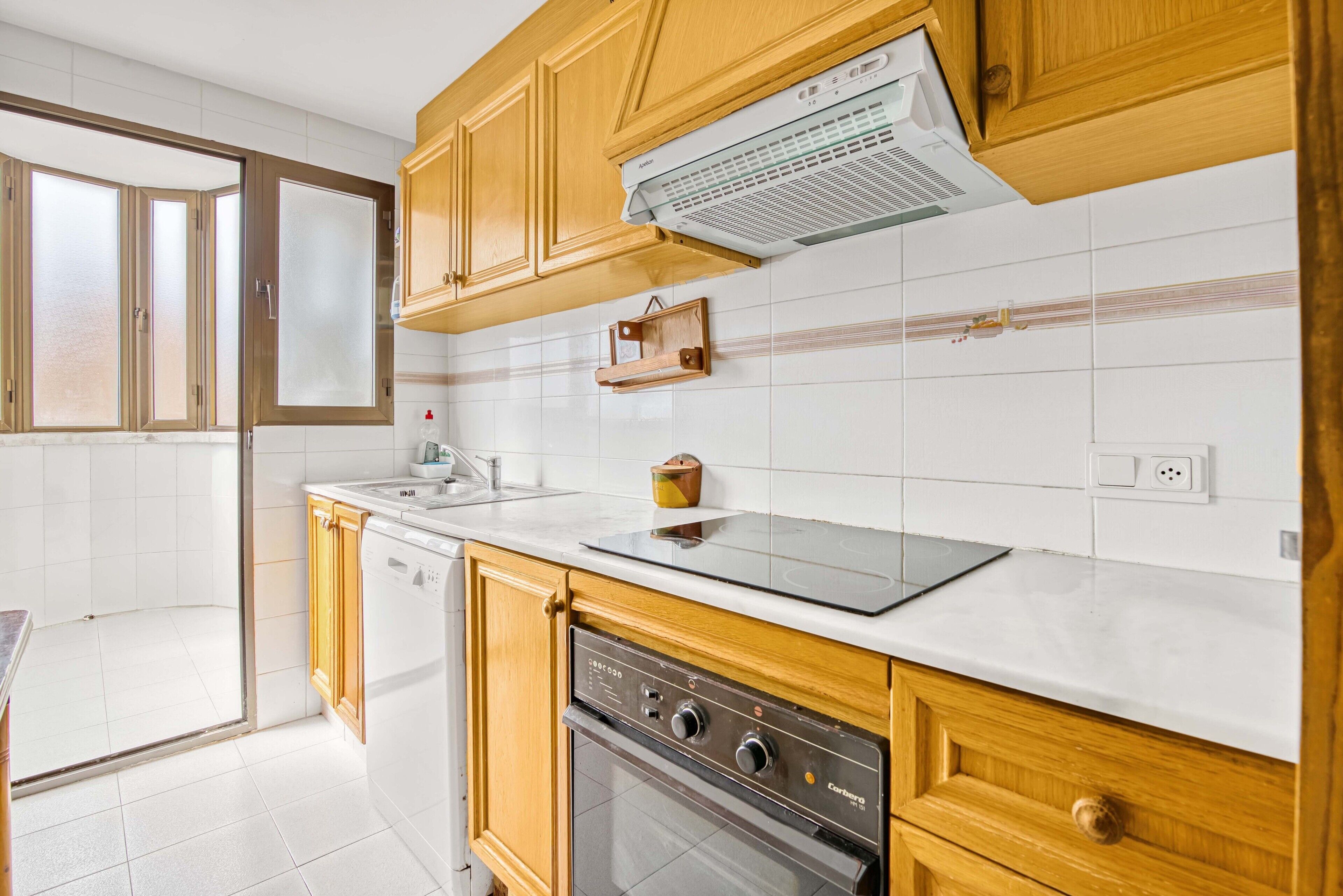Una cocina con armarios de madera, lavavajillas, horno, encimera con tetera y ventana.