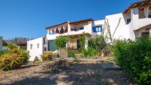 A white building with blue shutters, a garden with plants and a wooden structure, and a clear blue sky.