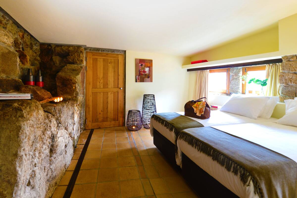 Una habitación con pared de piedra, puerta de madera, cama, mesita de noche y ventana con cortinas.