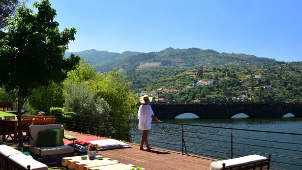 Una terraza de madera con mobiliario exterior, con vistas a un río y un puente, con vegetación y colinas al fondo.