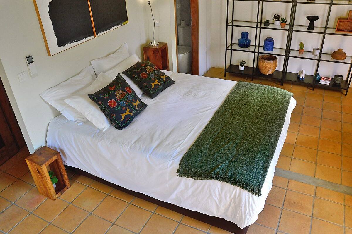 Una cama bien hecha con sábanas blancas y dos almohadas decorativas, un edredón verde y una mesita de noche de madera con una planta pequeña.