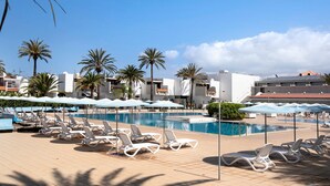Un espace piscine avec des chaises longues et des parasols, des palmiers et des bâtiments modernes en arrière-plan.
