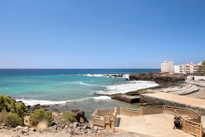 Vista costera con una terraza de madera, costa rocosa y edificios al fondo.