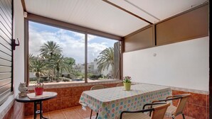 Une terrasse couverte avec une table et des chaises, un pot de fleurs et une vue sur des palmiers et des bâtiments.