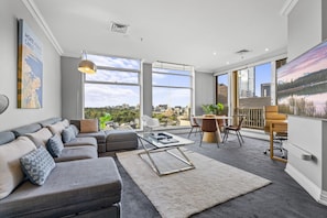 A modern living room with a sofa, coffee table, and dining area with chairs. Large windows offer a city view. - Bright, Luxury Penthouse with Amazing Views (Melbourne)