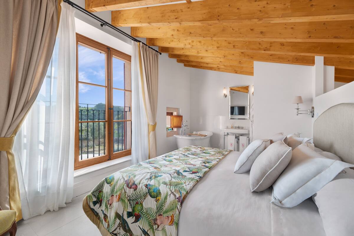Une chambre avec un grand lit, une fenêtre avec des rideaux, un plafond en bois, une salle de bain avec un lavabo et un miroir, et une vue sur la verdure à l’extérieur.