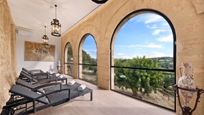 Une pièce baignée de lumière, avec des fauteuils en osier, une grande fenêtre en arc de cercle et une vue sur un vignoble.