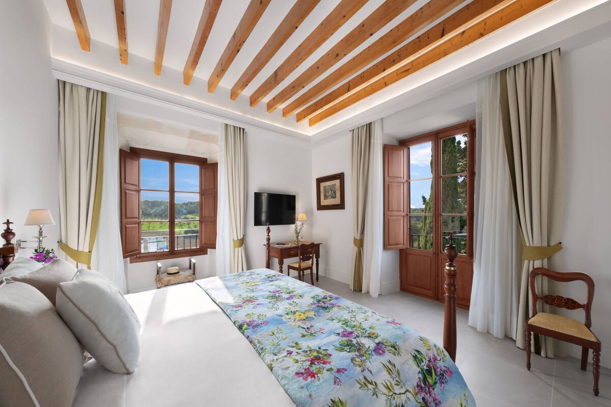 Une chambre à coucher comprenant un lit, un bureau, une chaise, un téléviseur et de grandes fenêtres avec des rideaux.