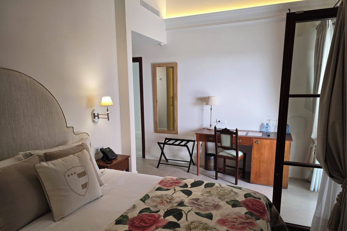 Une chambre d’hôtel avec un lit, un bureau, une chaise, une lampe et une salle de bain visible à travers une porte ouverte.