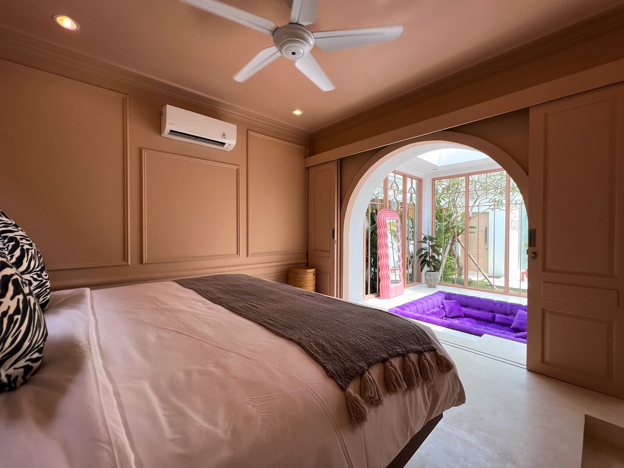 Ein Schlafzimmer mit einem Bett, einem Deckenventilator, einer Klimaanlage, einem Fenster und Blick auf einen violetten Sitzbereich draußen.