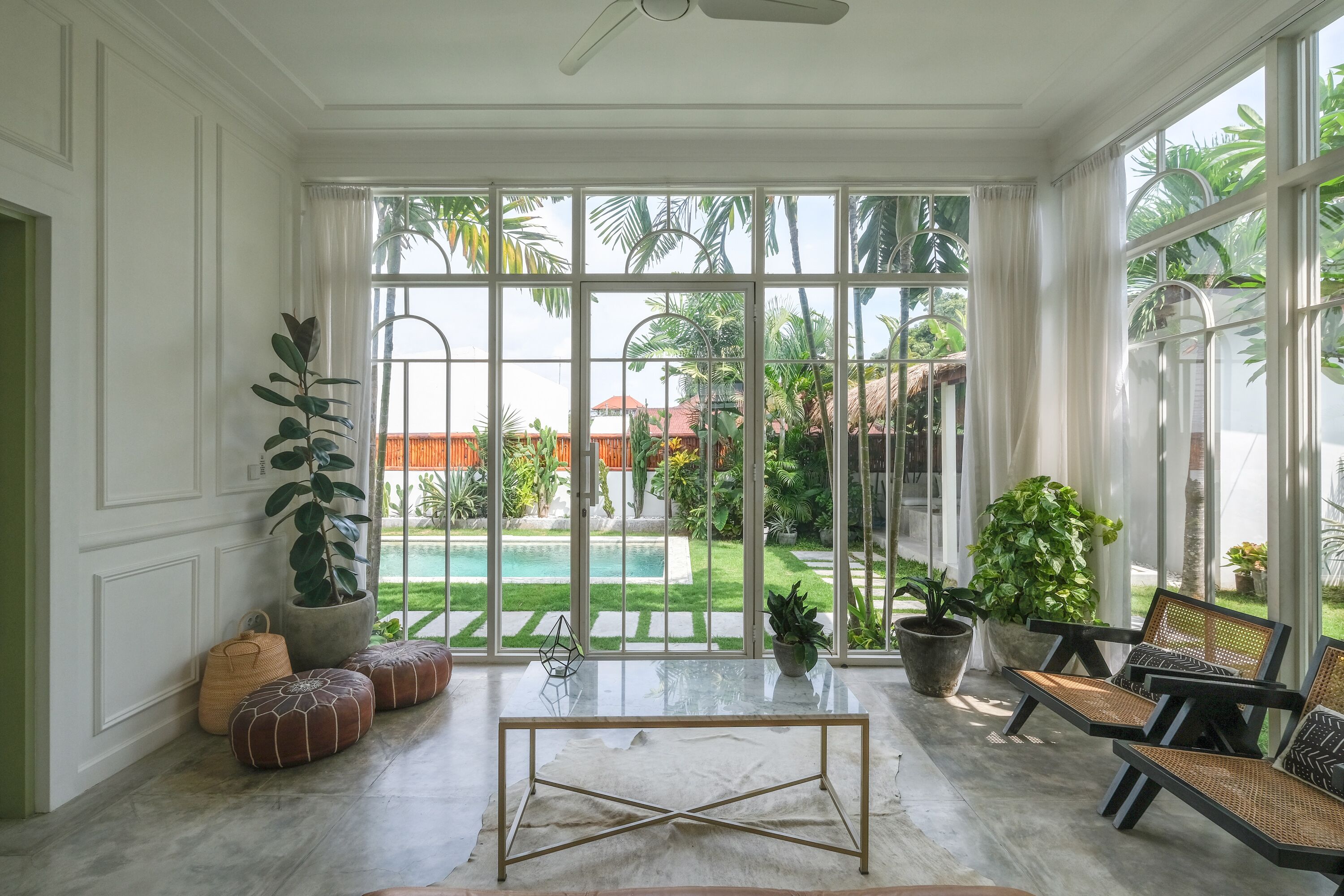 Amplia habitación con un gran ventanal que da a una piscina y jardín. Hay una mesa de vidrio con dos sillas y varias plantas en macetas.