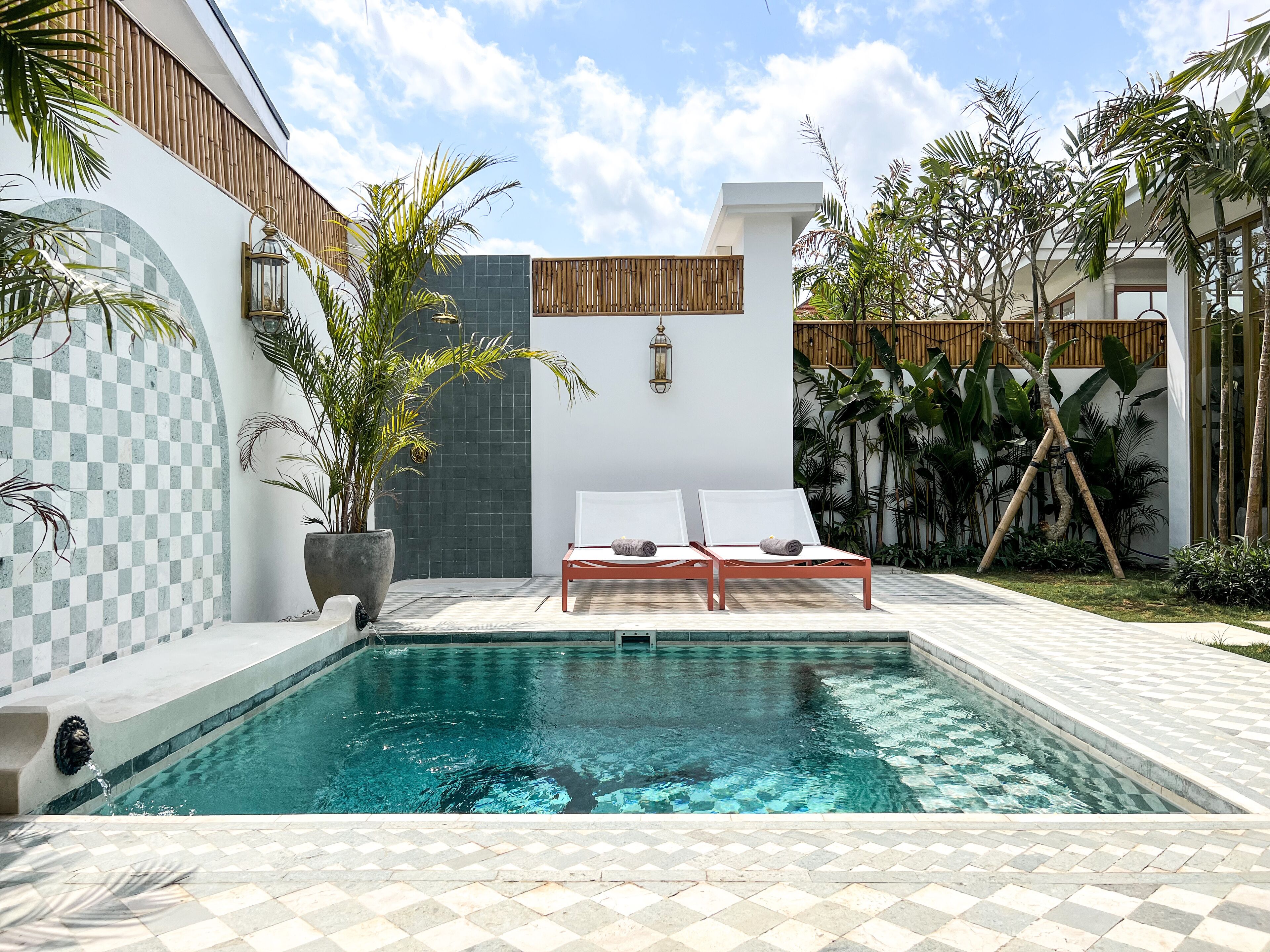 Una piscina con dos sillas de descanso, rodeada de plantas en macetas y un muro a cuadros.