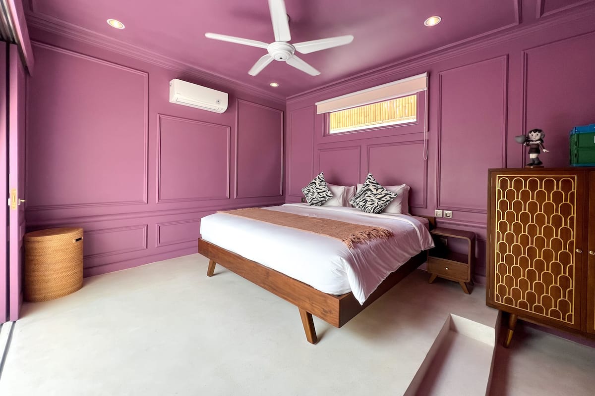 A bedroom with a bed, a ceiling fan, an air conditioning unit, a wooden cabinet, and a window with blinds.