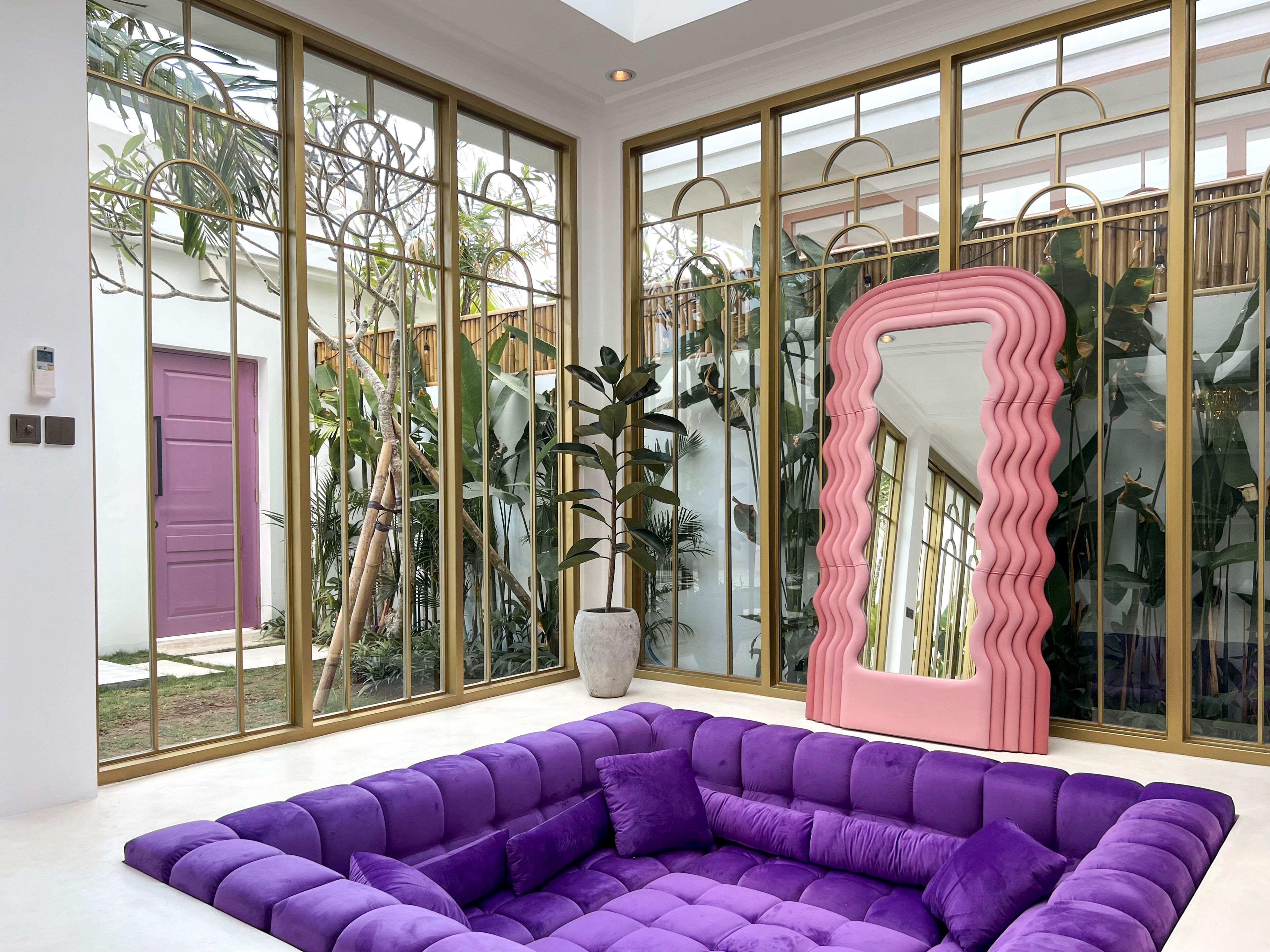 A modern living room with a large purple sectional sofa, a pink mirror, and large windows with a view of greenery outside.