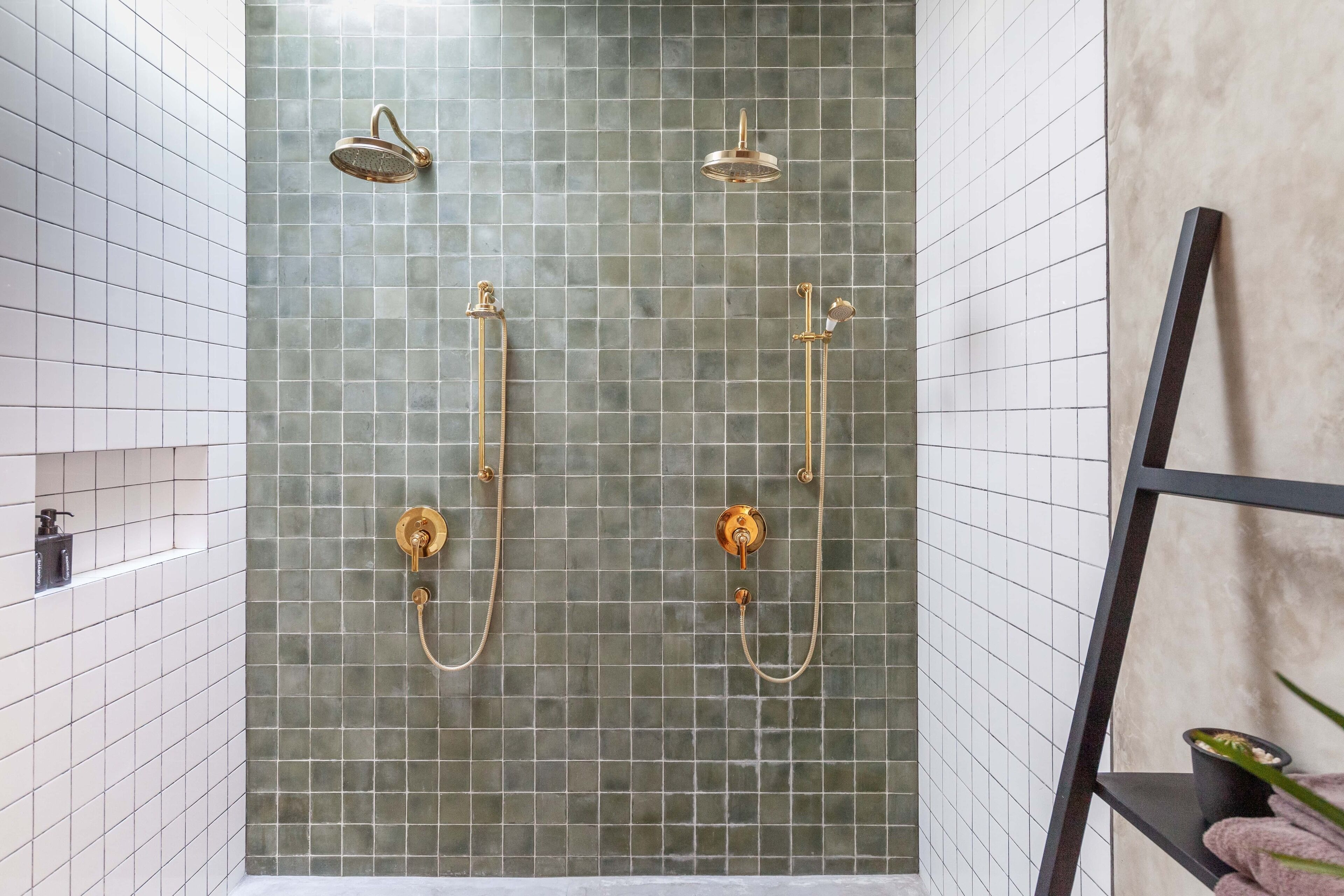 Un baño con pared de ducha en azulejos verdes, un estante negro con toallas y una pequeña planta.
