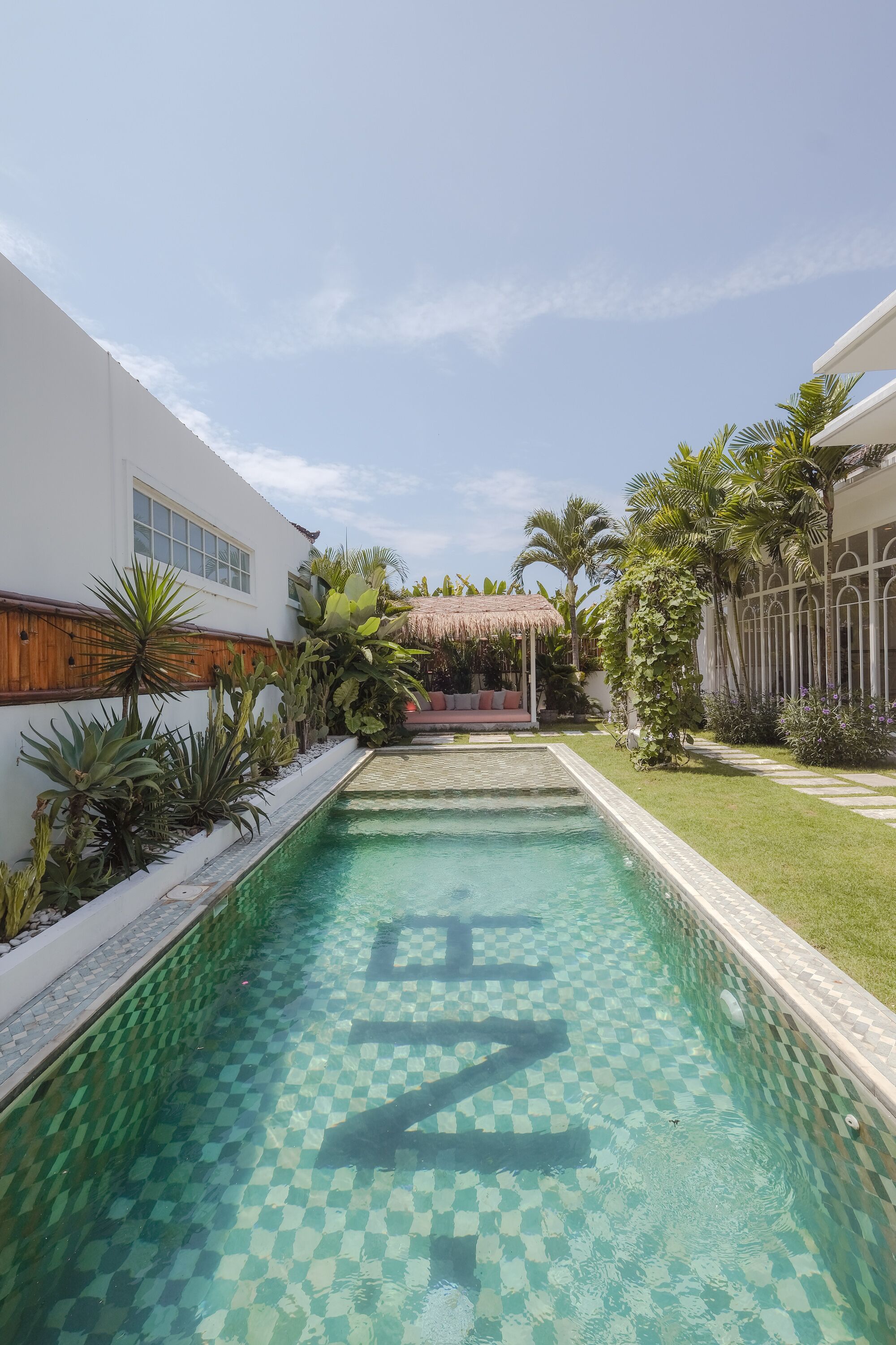 A swimming pool with a checkered pattern, surrounded by lush greenery and a thatched-roof structure.