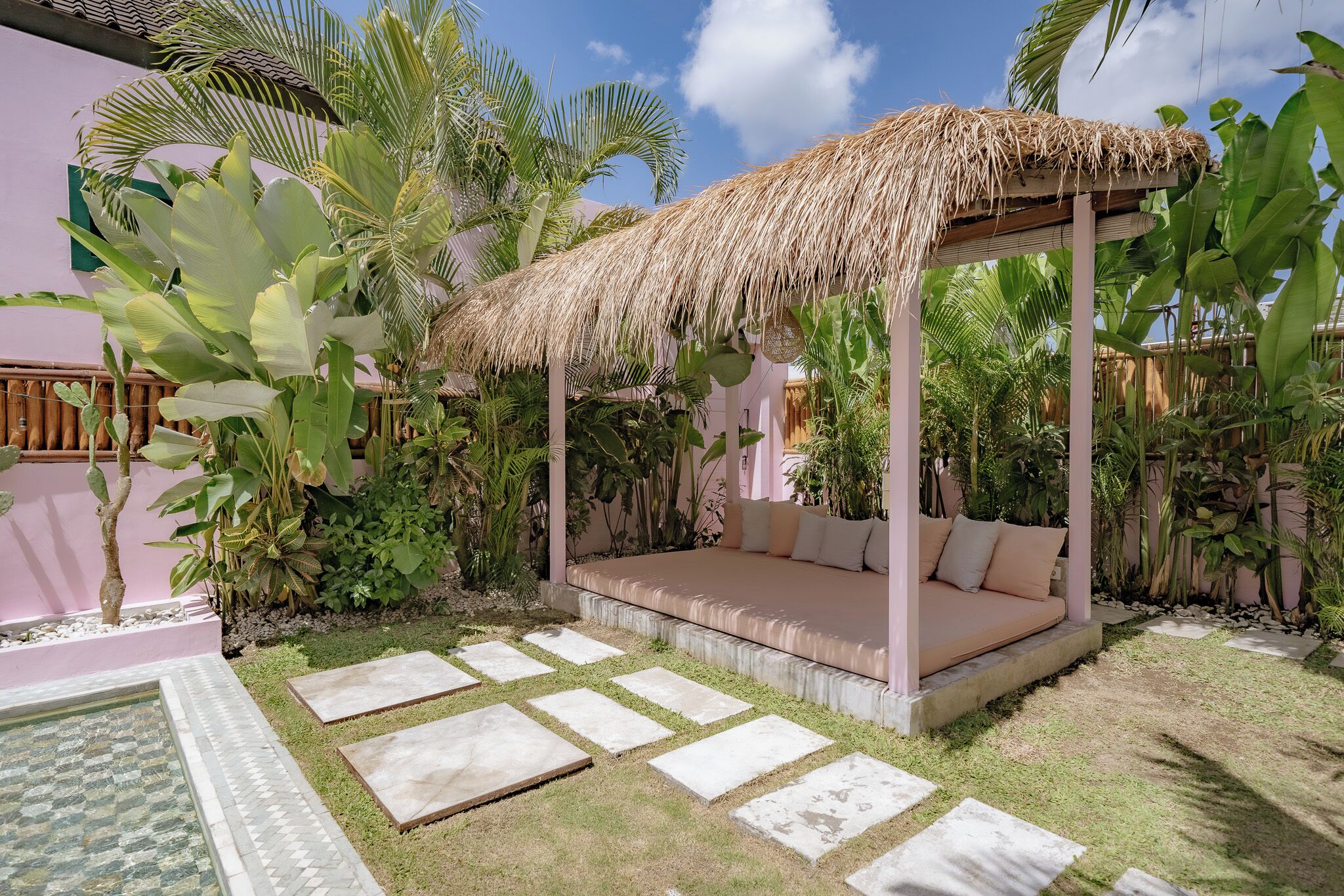 Un área de estar al aire libre con techo de paja, un banco acolchado y abundante vegetación tropical, todo ello rodeado por un sendero de piedra con diseño.