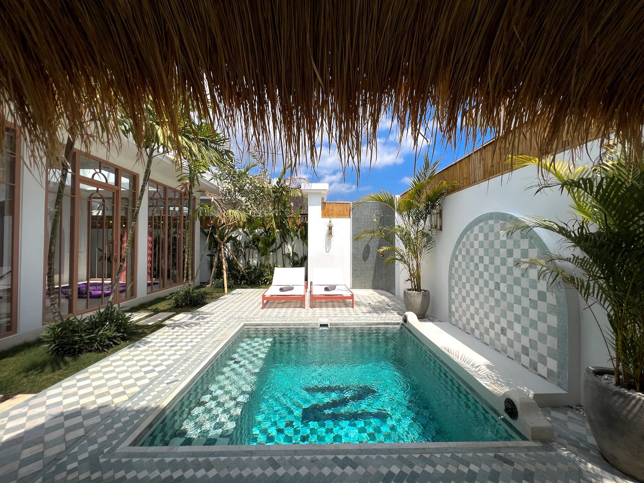 A swimming pool with a checkered tile pattern, surrounded by a thatched roof and potted plants.