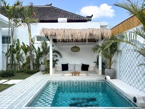 A pool area with a checkered tile pattern, a thatched roof seating area, and various tropical plants.