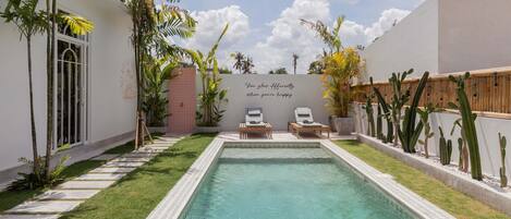 A swimming pool with two lounge chairs, surrounded by a garden with various plants and a wall with a motivational quote.