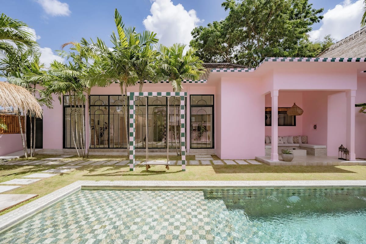 A pink building with a swimming pool, palm trees, and a thatched roof structure.