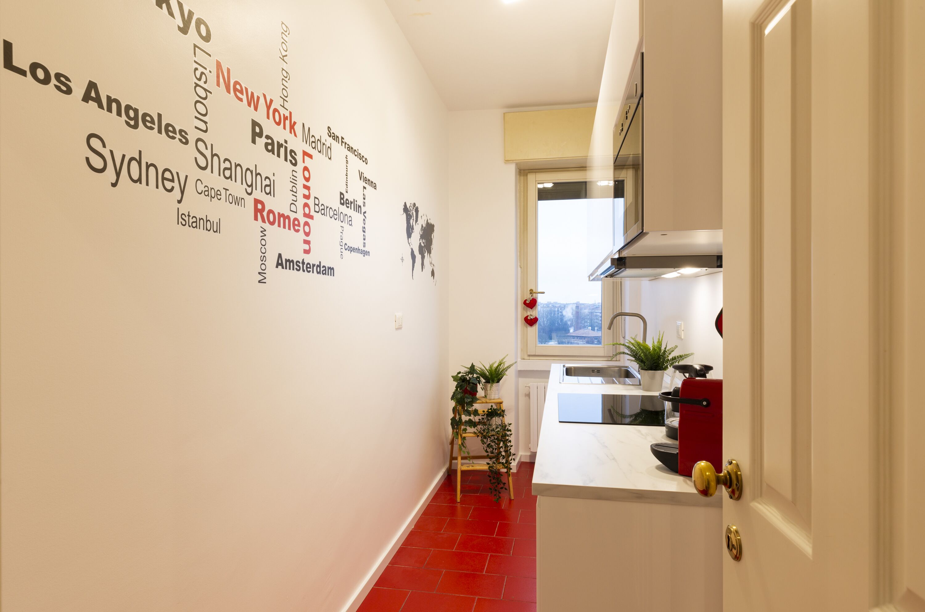 Een smalle gang met een rode betegelde vloer, een keuken met een spoelbak en fornuis, en een muur versierd met stadsnamen.