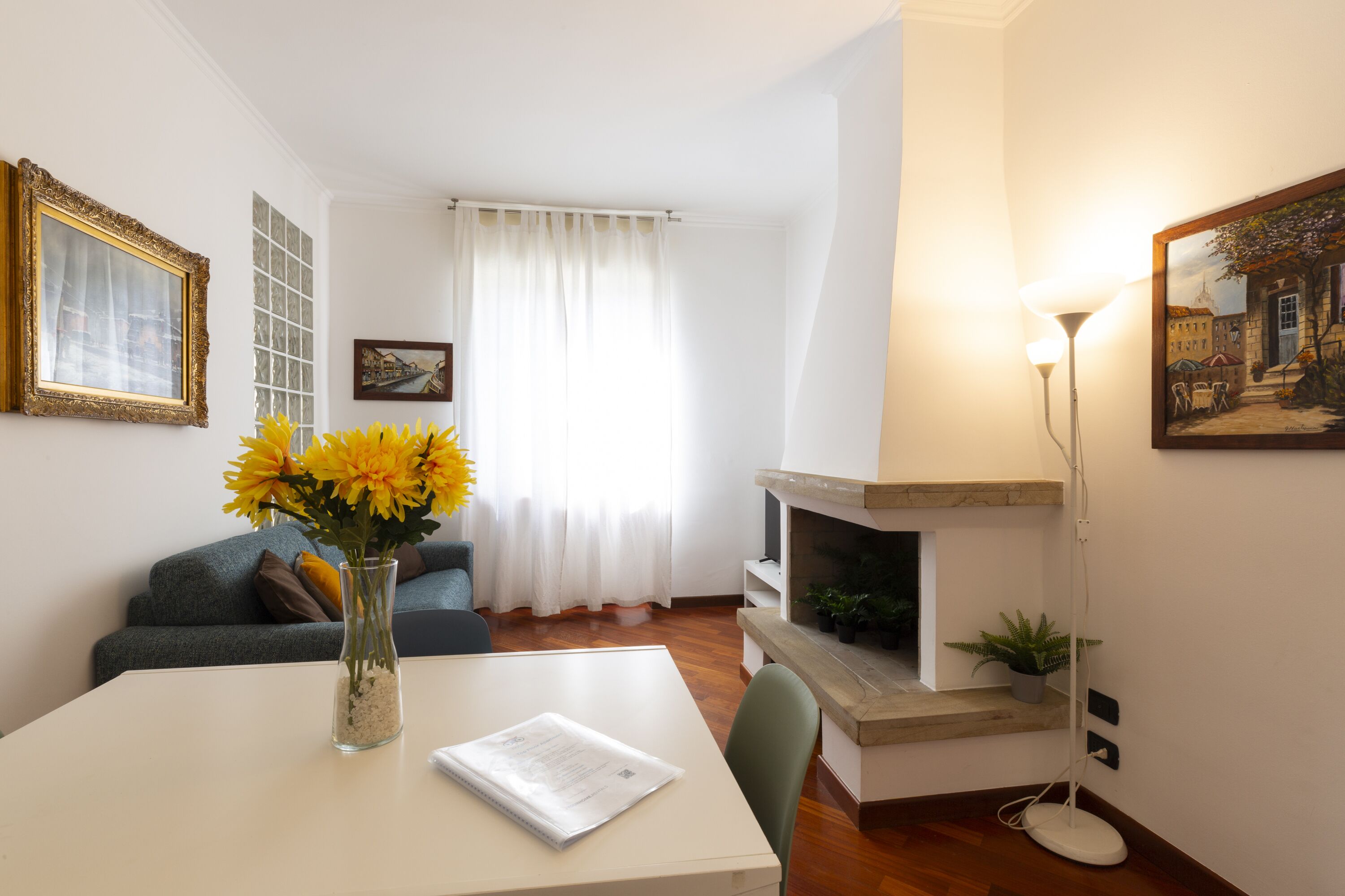 A living room with a white table, a vase with yellow flowers, a sofa, a fireplace, and a painting on the wall.