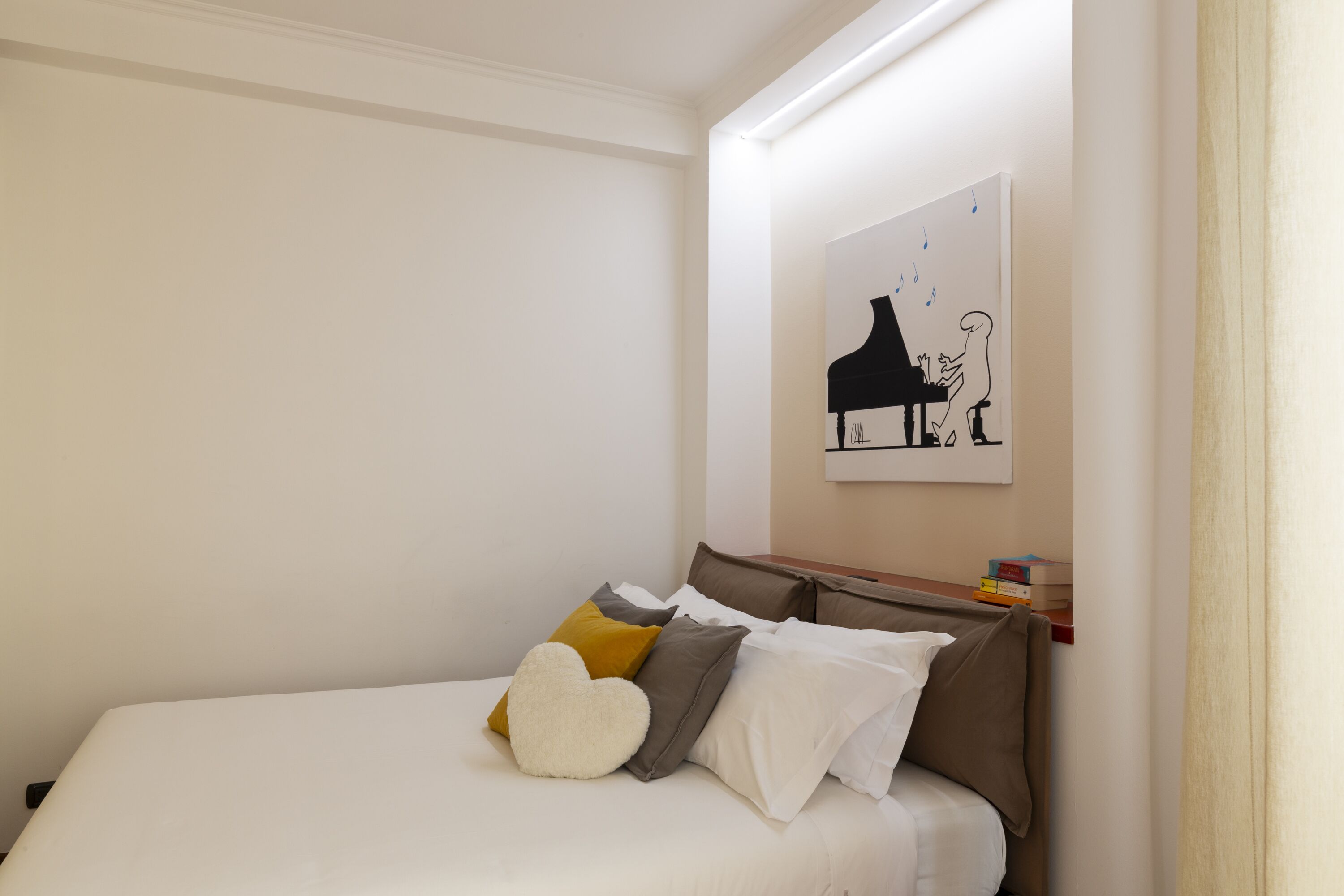 A neatly made bed with a heart-shaped pillow, a wooden nightstand with books, and a wall-mounted artwork of a piano.