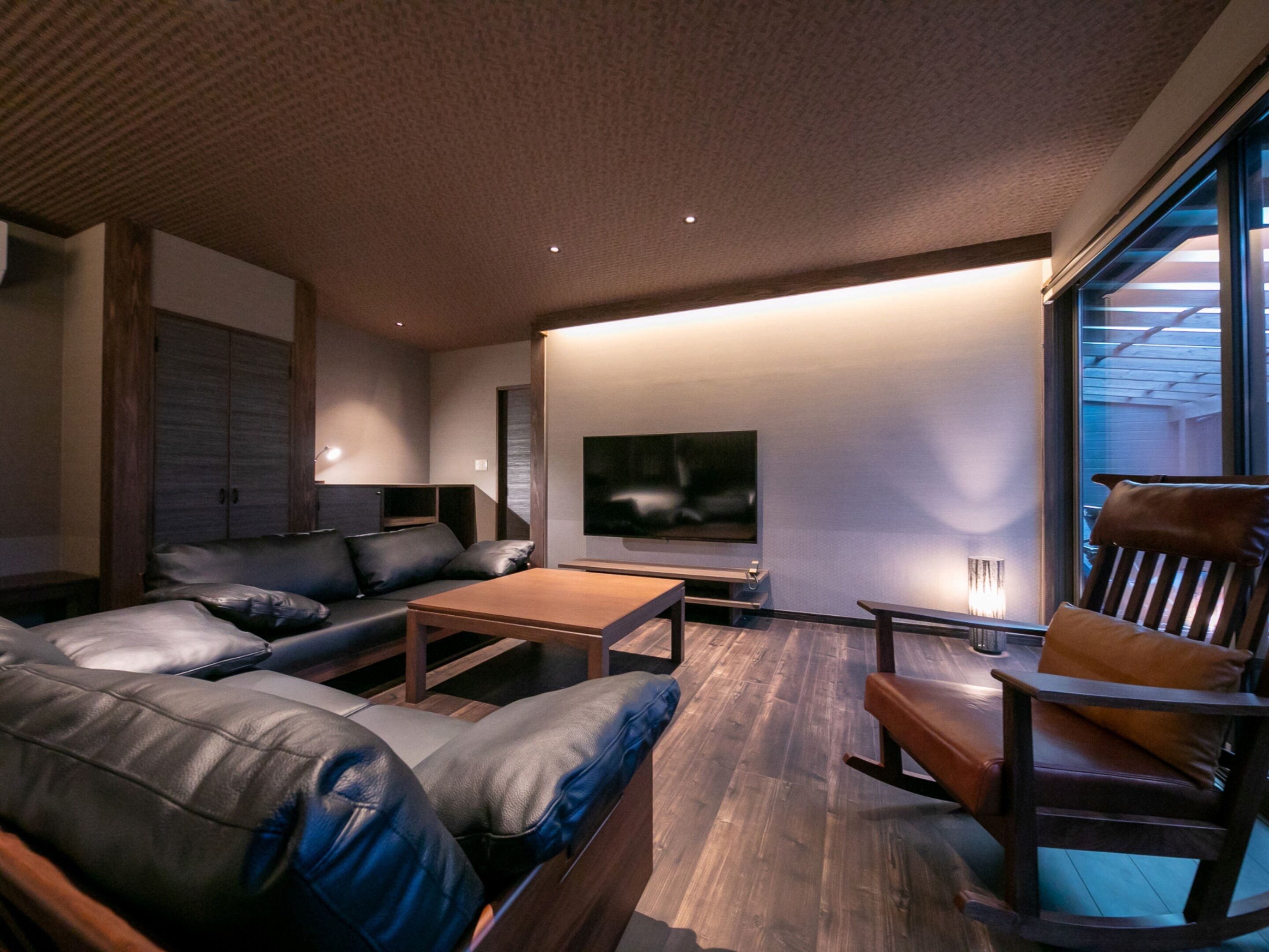 A modern living room with a sofa, coffee table, and a wooden rocking chair. There is a flat-screen TV mounted on the wall and a window providing natural light.