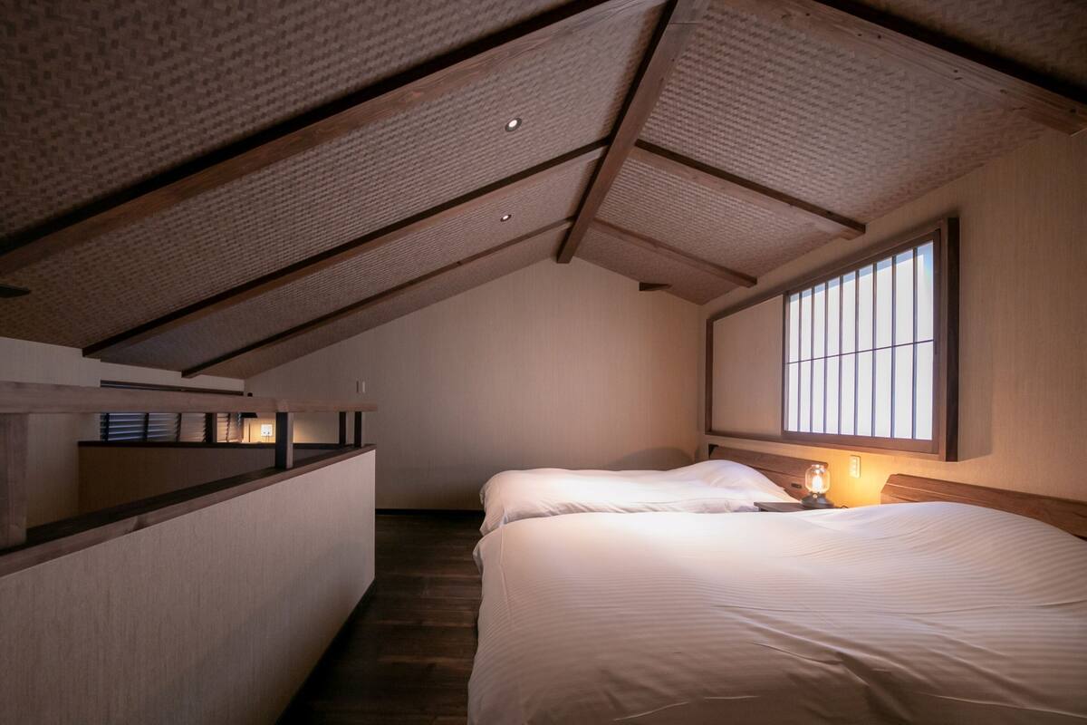Quarto com teto inclinado, uma cama, mesa de cabeceira e janela com persianas.