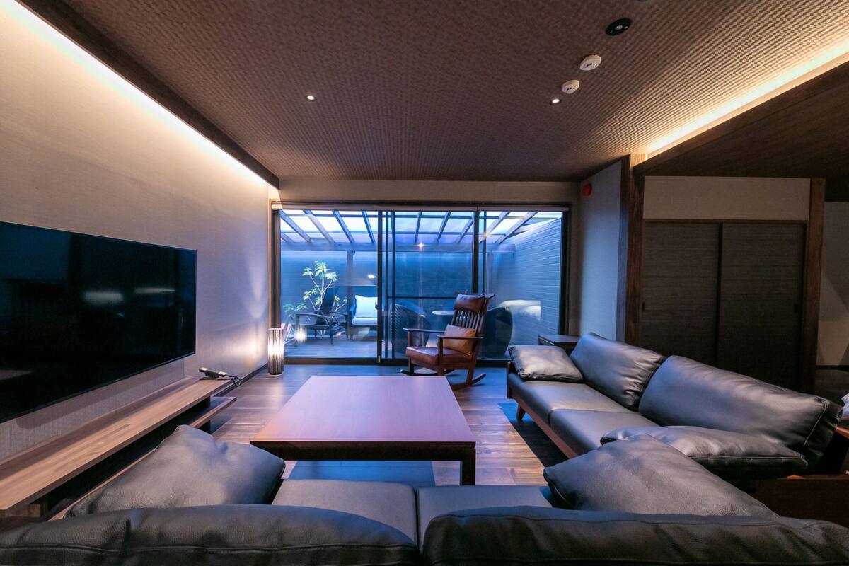 Uma sala de estar moderna com uma TV de tela plana, sofás de couro, uma mesa de centro de madeira e uma porta de vidro que dá para uma área externa com uma cadeira e uma mesa.