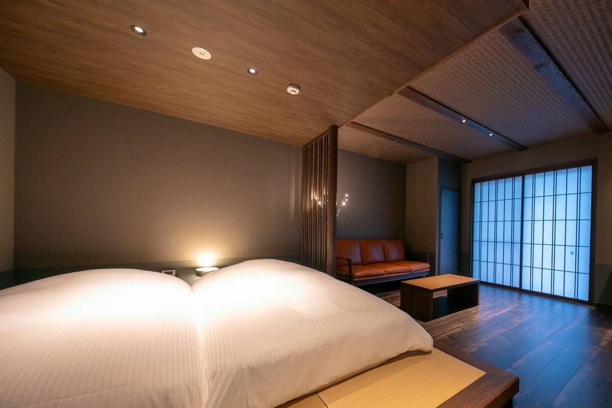 Quarto de hotel moderno com uma cama grande, um pequeno sofá, piso de madeira e uma janela ampla com vista.