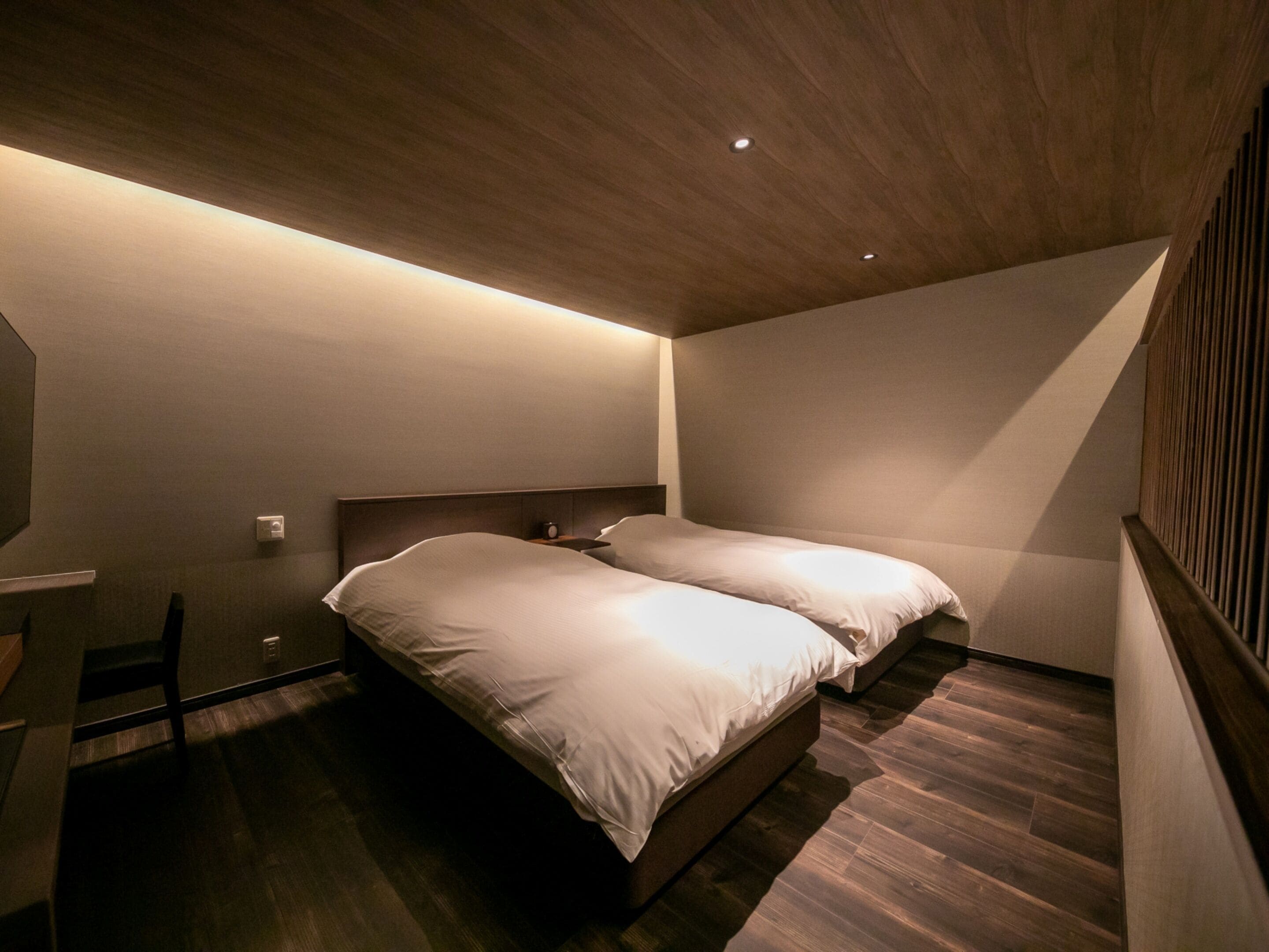 A small, dimly lit room with a single bed, a desk, and a chair.