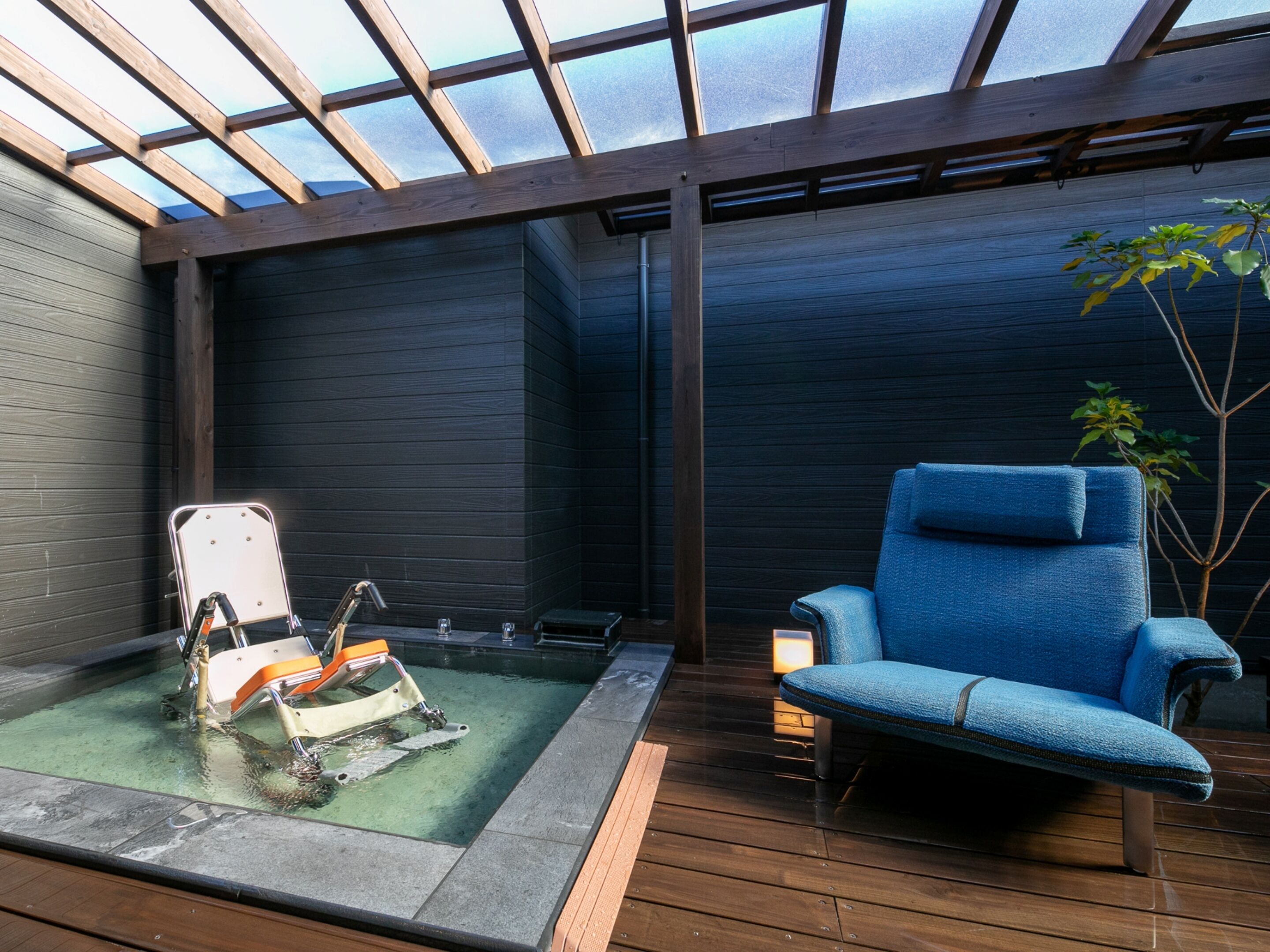 A spa area with a wooden deck, a blue chair, a massage table, and a glass-enclosed room with a view.