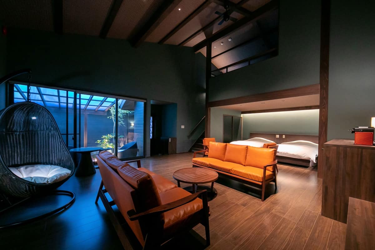 Quarto de hotel moderno com uma cama grande, um sofá, uma cadeira e uma pequena mesa. Tem uma varanda com vista para o exterior.