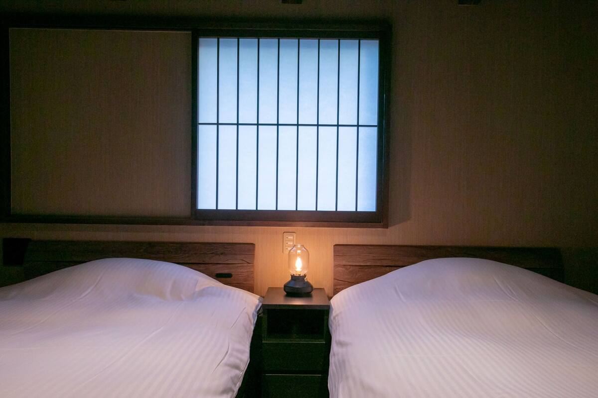 Quarto com duas camas, uma mesa de cabeceira com abajur e uma janela com persianas verticais.