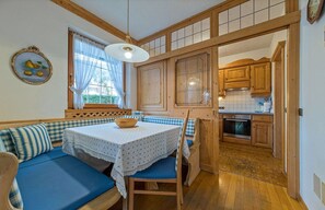 Un comedor con una mesa y un banco tapizado en azul, una ventana con cortinas, y una cocina visible a través de una puerta abierta.