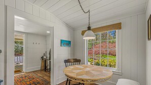 Una habitación con paredes blancas empañadas, una mesa redonda con mantel tejido, una lámpara colgante y un ventanal con vistas a un jardín.