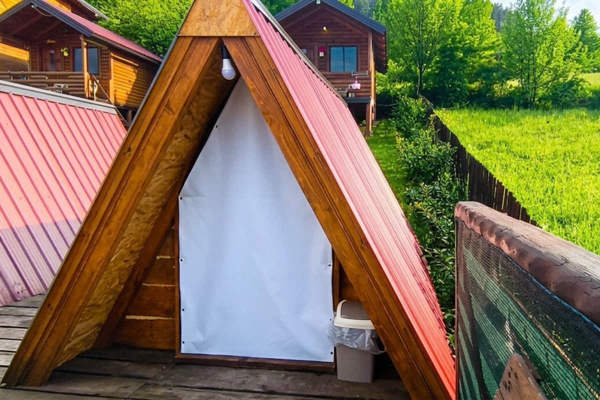 Eine hölzerne, dreieckige Konstruktion mit weißem Innenraum, neben einer Holzterrasse und einem grünen Feld.