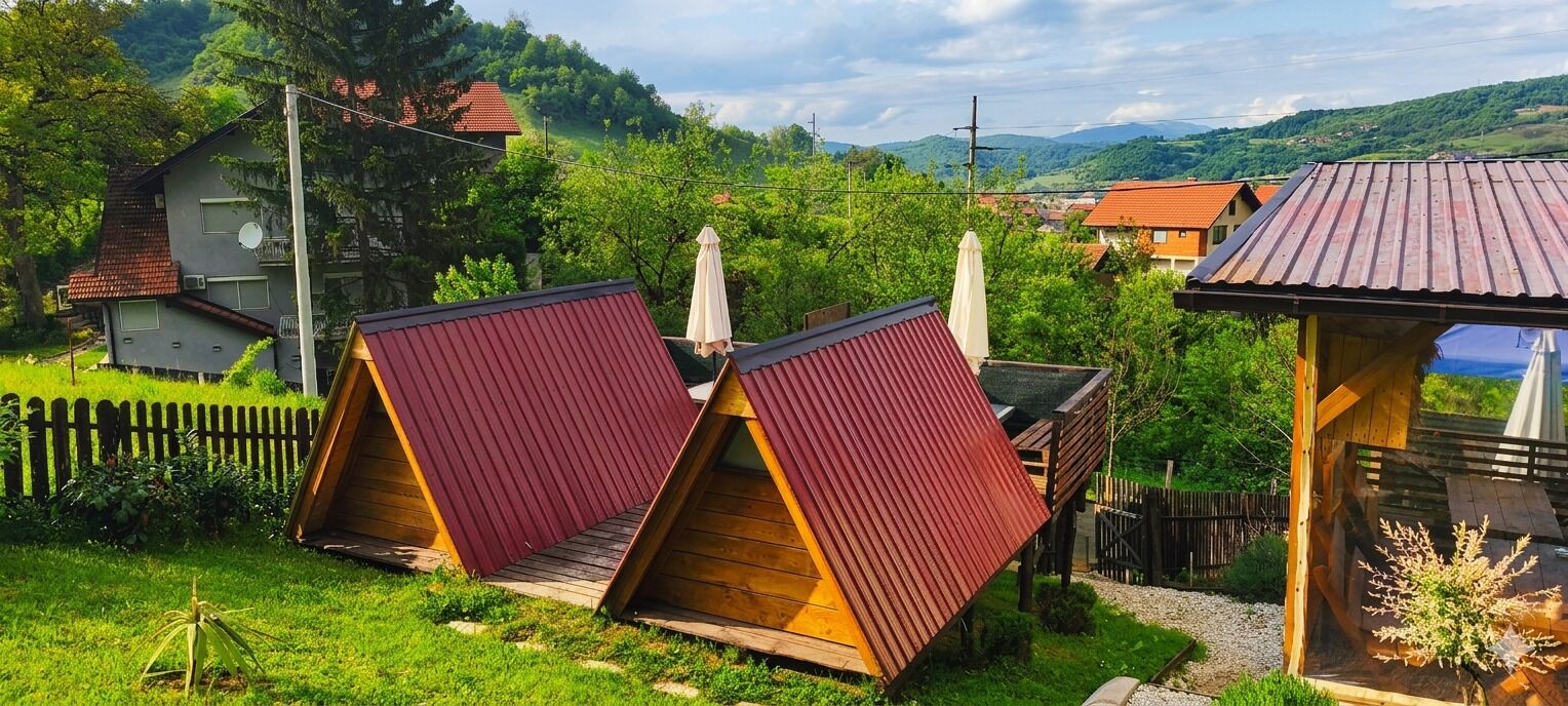 Un paysage rural avec deux cabanes en bois aux toits rouges, entourées de verdure et d’une clôture en bois.