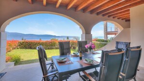 A covered outdoor dining area with a table set for two, overlooking a scenic view of the sea and mountains.