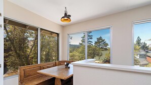 Una habitación con un banco de madera, una mesa y ventanas amplias que ofrecen vistas a árboles y un edificio lejano.