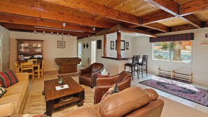 A cozy living room with wooden beams, a fireplace, leather sofas, and a dining area with a wooden table and chairs. - Alpine Skies by Destination Big Bear : Peaceful Setting in Moonridge! Walk to Bear Mtn and the National Forest! Hot Tub! (Big Bear Lake)